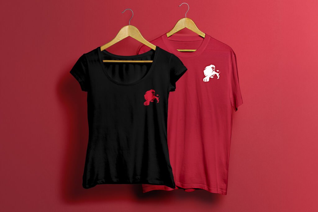 Two designer t-shirts on wooden hangers against a red wall: a black fitted shirt with a red lion emblem on the left chest and a red unisex shirt with a white lion emblem in the same spot. - FelipeIgiesias.com