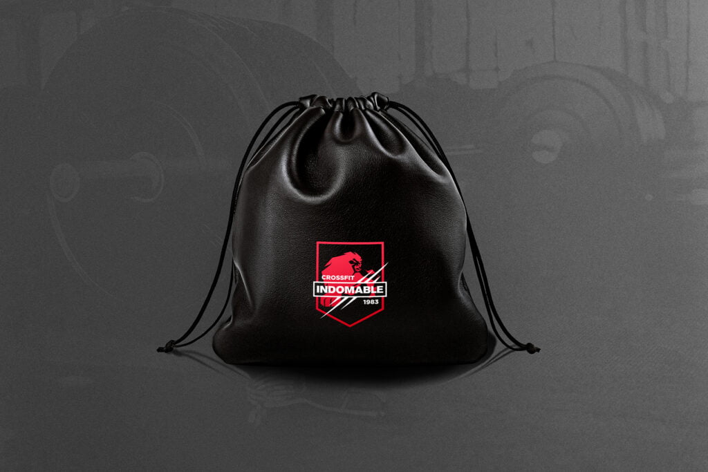 A black drawstring bag with a striking red and white CrossFit Indomable lion logo showcases bold branding, set against a dark, blurred background filled with gym equipment. - FelipeIgiesias.com