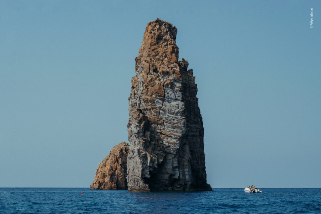 "Towering volcanic rock formation rising from the sea, with a small white boat to the right and a clear blue sky above – photography by Felipe Iglesias.
