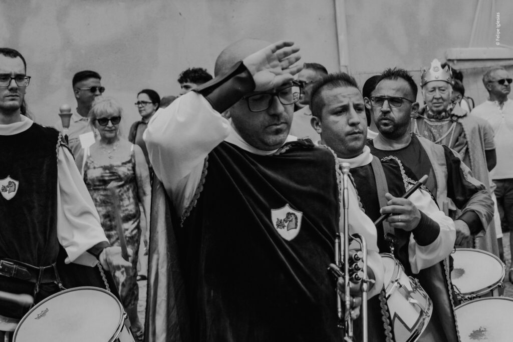 An overheated, bald trumpeter wearing glasses wipes his forehead while the rest of the band continues to play in vibrant medieval attire, leading a colourful procession followed by a large crowd at Ntinna a Mari, Cefalù– picture by Felipe Iglesias.