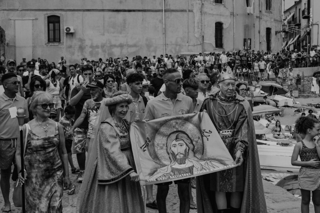 The 'King and Queen' of Cefalù, accompanied by the 2023 'Ntinna a Mari' winner, carry the flag featuring a Byzantine portrait of Christ Pantocrator through the town, followed by a crowd of curious onlookers during the traditional celebration – photography by Felipe Iglesias.