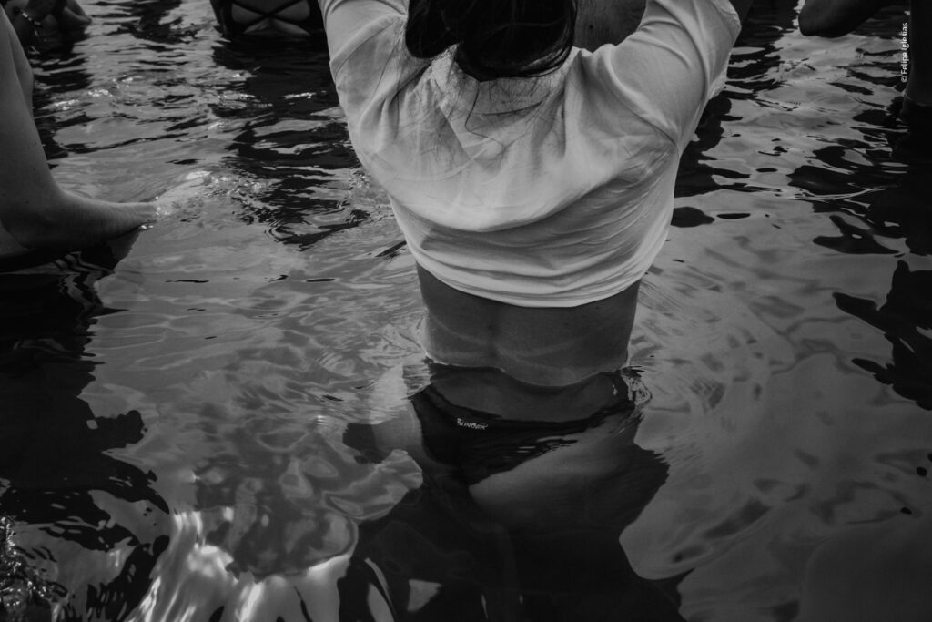 A young woman carefully wades into the mossy waters during the 2024 'Ntinna a Mari' event in Cefalù, lifting her t-shirt to stay dry, with the festive atmosphere of the traditional coastal celebration in the background – photography by Felipe Iglesias.