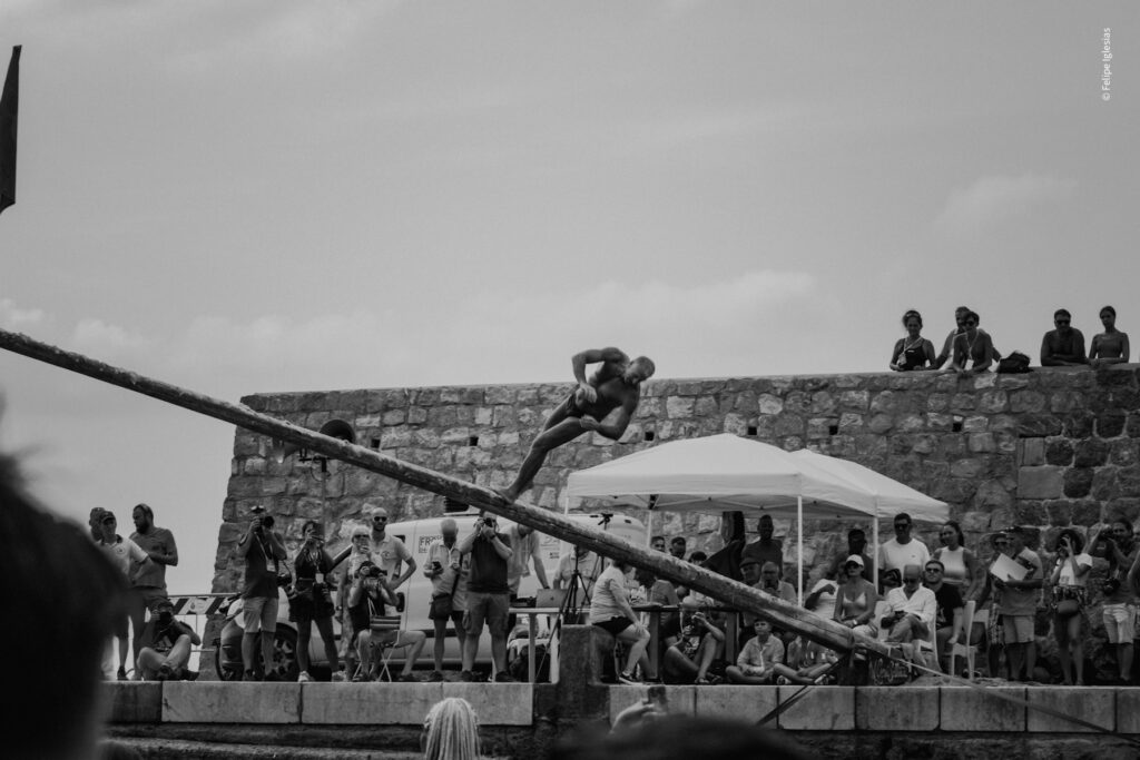 A contestant slips and falls from the greased pole during the 2024 'Ntinna a Mari' competition in Cefalù, capturing the thrill and challenge of this traditional seaside event – photography by Felipe Iglesias.
