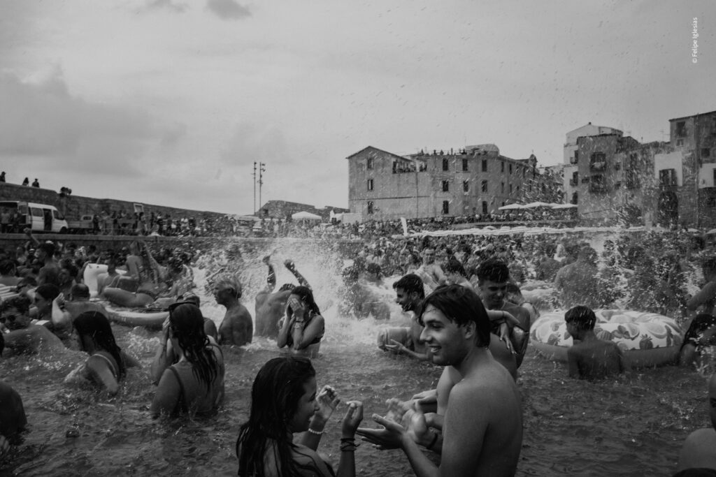 "Festive celebration of 'Ntinna a Mari' in Cefalù, with crowds gathered at the harbour and people joyfully splashing in the sea during the traditional coastal event – photography by Felipe Iglesias.