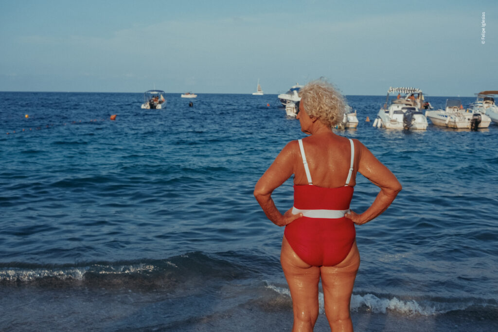 Tanned elderly woman with white curly hair, wearing a red swimsuit with a horizontal white stripe at the waist, stands with her feet in the water, gazing out at the Aeolian Islands Sea, with gentle waves and several white boats in the deep blue background – photography by Felipe Iglesias.