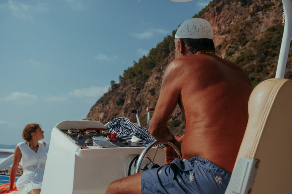 Deeply tanned skipper, half-naked and wearing blue shorts, a pirate-style earring, and a traditional white Aeolian cap, steering the boat while gazing towards a rocky cliff, with a woman in a white dress visible in the background – photography by Felipe Iglesias.