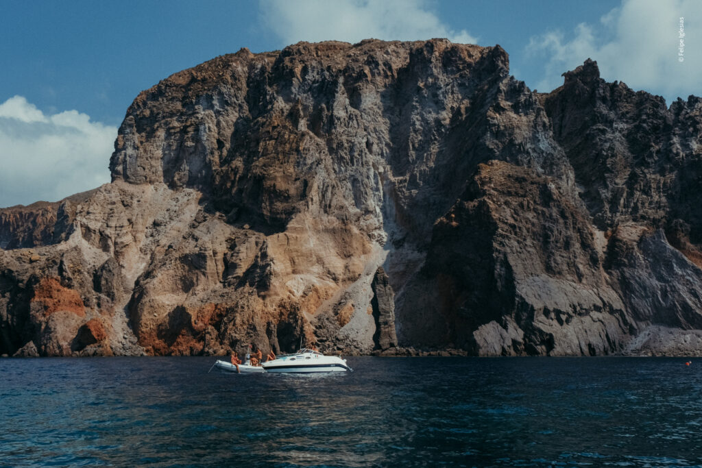 A small boat with a tender floats near the base of a rugged volcanic cliff, where several half-naked people are relaxing, set against the deep blue Aeolian Sea and a sky dotted with sparse white clouds – photography by Felipe Iglesias.