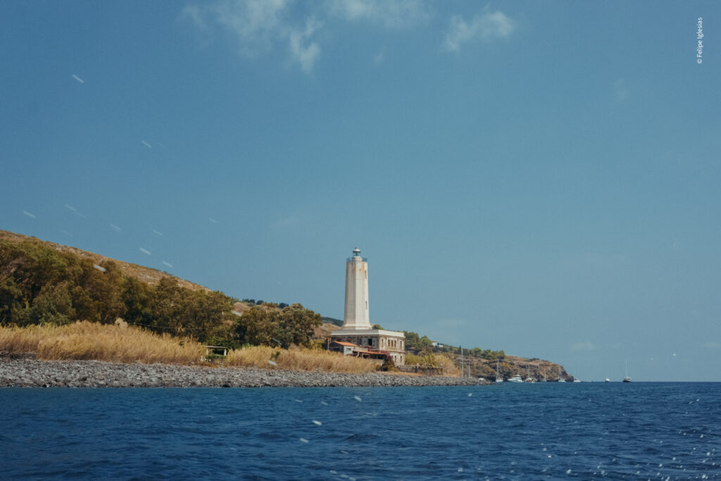 An old lighthouse stands prominently at the edge of Lipari Island’s rugged coastline, perched above a bushy slope, with a clear blue sky and deep blue sea stretching into the distance, where a few boats can be seen – photography by Felipe Iglesias.