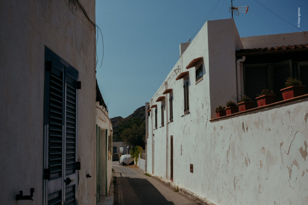 Eerie and solitary narrow street on Vulcano Island, lined with traditional white Aeolian houses; the larger house on the right features four upper-floor windows and a ground-floor door facing the street, with a covered car parked nearby and a partial view of a hill in the background – photography by Felipe Iglesias.