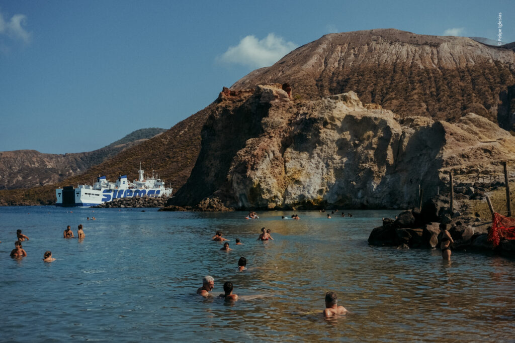 Partial view of Vulcano’s harbour from Hot Waters Beach, featuring the volcanic crater in the background, a large Siremar ferry docked near the shore, and several people relaxing in the warm, shallow waters in the foreground – photography by Felipe Iglesias.