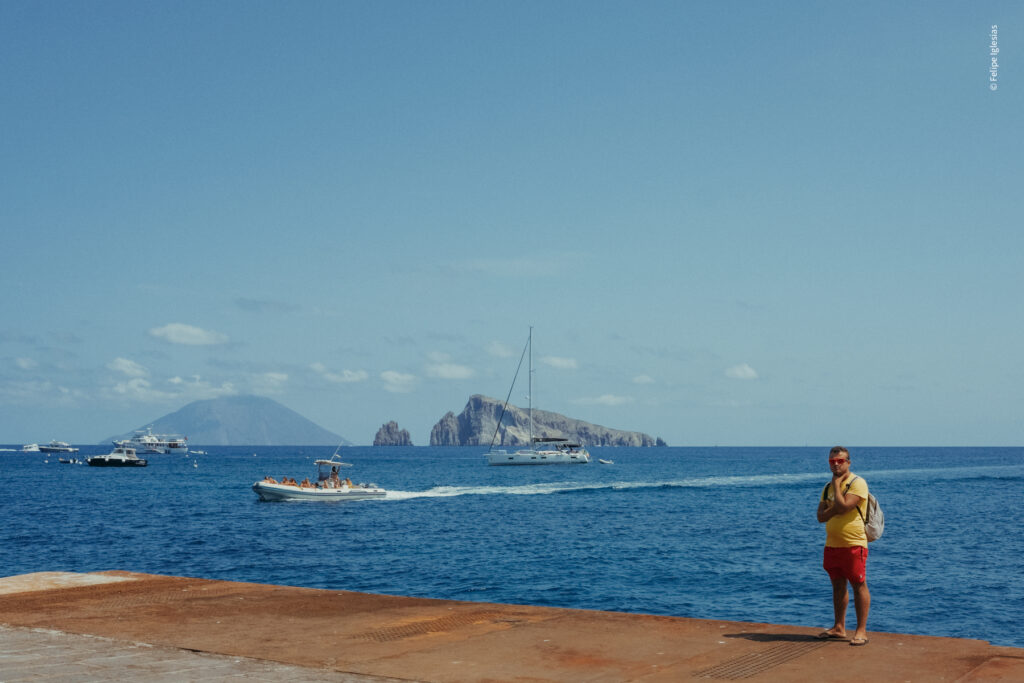 Scenic view of Panarea’s harbour with Basiluzzo Island at the centre and Stromboli Island on the left, several boats filled with people near the shore, and in the foreground, a man wearing a yellow T-shirt, red shorts, sunglasses, sandals, and a grey backpack looking directly at the camera – photography by Felipe Iglesias.