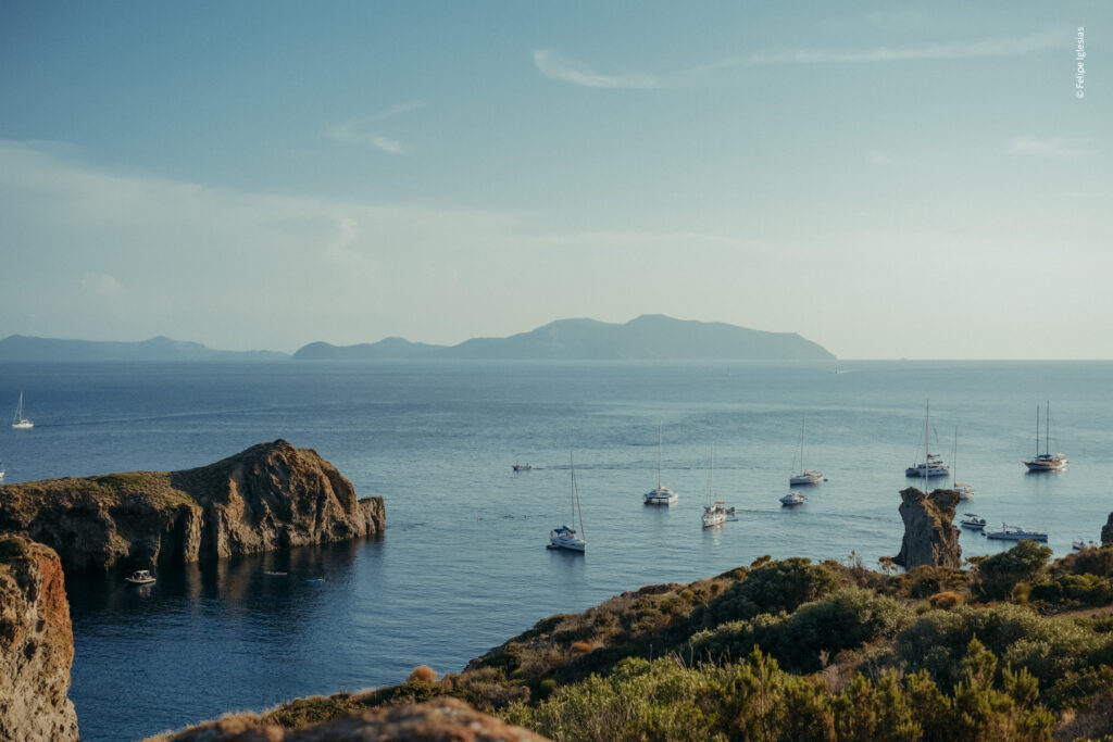 Breathtaking sunset view over the sea at Cala Junco, Panarea Island, featuring 12 white boats floating on deep blue waters, with Lipari Island in the background – photography by Felipe Iglesias.