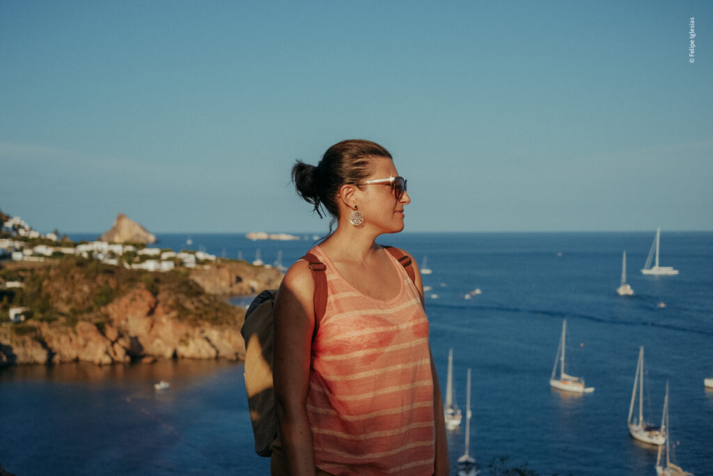 Beautiful Sicilian woman wearing sunglasses, statement earrings, an off-shoulder orange striped top, and an ochre backpack, gazing at the Aeolian Sea from a scenic summit with a deep blue sky and Mediterranean coast landscape in the background – photo by Felipe Iglesias.
