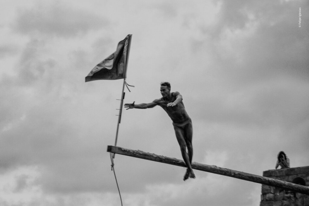 The exact moment Antonino Papa triumphs in the 2024 edition of the 'Ntinna a Mari' contest in Cefalù, leaping to seize the flag at the end of the greased pole beneath a dramatic, cloudy sky – photography by Felipe Iglesias.